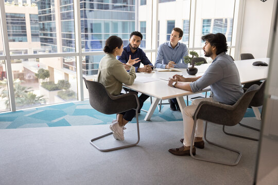 Diverse team of young male colleagues listening to speaking Indian business leader at meeting table in collaboration room interior, talking in contemporary workspace. Candid shot with copy space - Powered by Adobe
