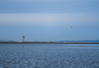 Two airplanes are seen taking off over the bay near San Francisco International Airport at dusk, with control towers and city lights visible in the distance.