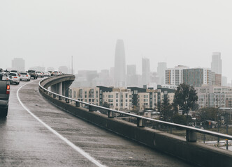 Obraz premium San Francisco freeway with heavy traffic on a rainy day. The wet road curves towards the downtown skyline, featuring high-rise buildings and misty urban architecture.