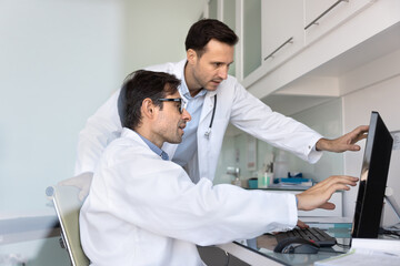 Two medical professionals meeting and talking at work computer in clinic office, touching and pointing at monitor, discussing patients case, diagnosis, examination electronic result