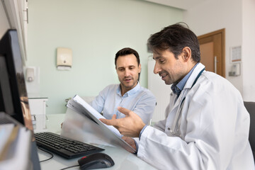 Obraz premium Positive middle aged physician man giving consultation to patient, reading paper history, analyzing medical records, explaining diagnosis, treatment, prescription, pointing at file, speaking, smiling