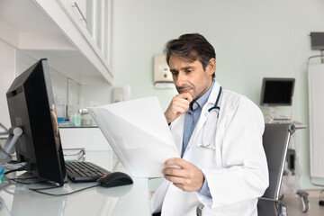 Focused Latin doctor man reading document carefully, reviewing paper medical report at workplace...