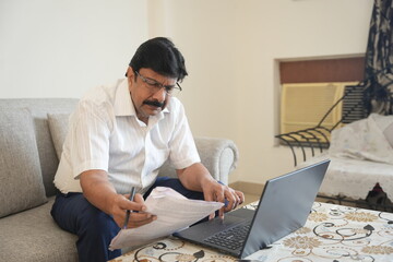 mature indian man working on laptop and checking document in a calm home setting. businessman and...