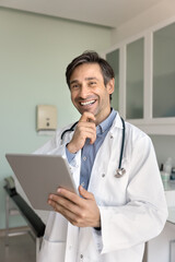 Cheerful Hispanic male general practitioner man using tablet in clinic office, analyzing medical records, touching chin, looking away, smiling, laughing, thinking, enjoying job in medicine