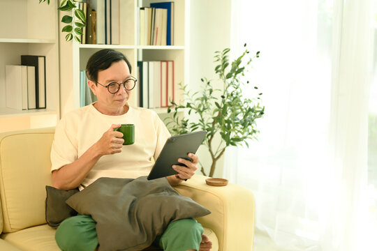 Happy senior man sipping coffee from a green mug and browsing on digital tablet - Powered by Adobe