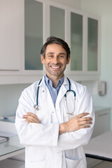 Positive Hispanic male doctor standing with hands folded, looking at camera with toothy smile, posing for vertical portrait in hospital lap, office, examination room alone, looking at camera