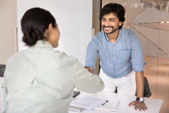 Happy confident young Indian businessman shaking hands with female business partner, colleague after signing contract, agreement, successful negotiation, giving handshake over table