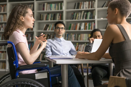 Social inclusion of people with disabilities. Teenage girl student wheelchair user lead group discussion seminar express idea to interested diverse multiracial mates on round table meeting at library