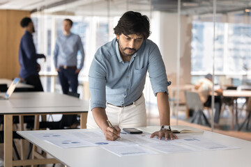Serious Indian business project leader man working on statistic reports, writing notes in...