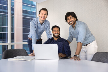 Casual portrait of happy male business colleagues meeting at laptop for teamwork, cooperation, having fun, laughing, smiling, looking away, sitting and standing at large office table