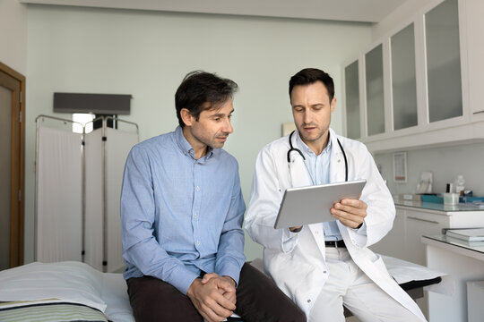 Serious doctor showing electronic checkup results to patient, using tablet computer, talking to man, explaining medical report, diagnosis after examination in clinic office - Powered by Adobe