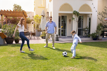 Happy young parents and son kid kicking soccer ball on grass at house, playing football outside...