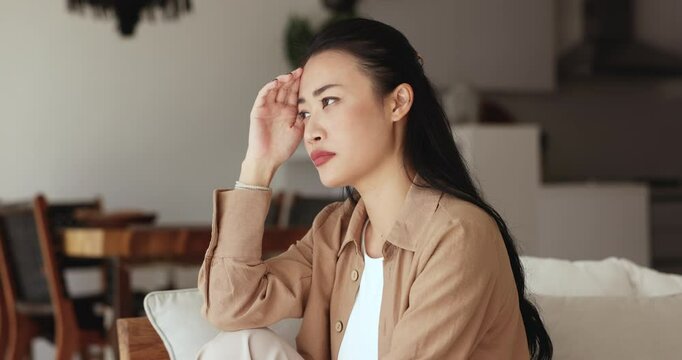 Depressed serious young Asian woman touching head and looking away, sitting on home couch, thinking on serious problems, troubles, hard decision, bad news, closing eyes hopelessly