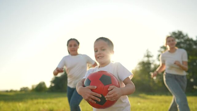 Happy family playing soccer in park. Smiling boy holds ball while running with family in park. Kids and mother enjoy active play in nature. Happy family bonding, outdoor fun concept in sunny park.
