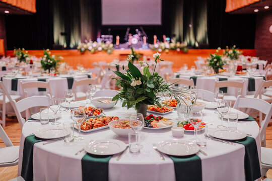 Close-up of an elegant round table setup with a floral centerpiece, plates, glasses, and food dishes in a banquet hall with a stage.