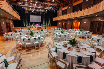 Large event hall with elegantly set round tables, floral centerpieces, food, and a stage prepared...