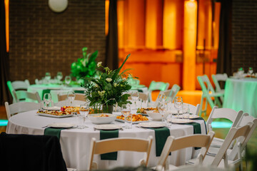 Elegant banquet table setup with white tablecloth, green accents, floral centerpiece, and dishes, in a warmly lit event venue..