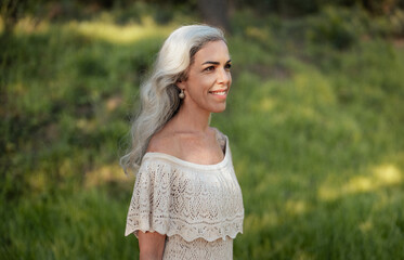 A graceful woman with long gray hair smiles warmly while standing in a sunlit outdoor environment....