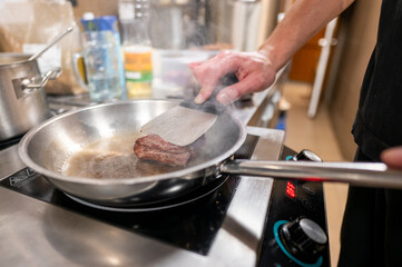 A chef skillfully flips a piece of meat in a stainless steel pan on a modern stovetop. The kitchen setting features various cooking tools and a hint of steam, showcasing culinary preparation.