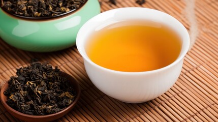 A cup of warm tea beside dried tea leaves on a bamboo mat.