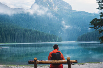 Male traveler with backpack in bright orange jacket on background of Black Lake, Durmitor Park...