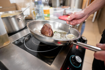 A chef sautéing a piece of meat in a frying pan on a modern induction cooktop. The kitchen...