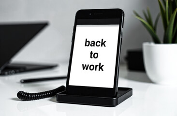 the inscription in black letters on the white screen of the phone is "Back to work". The phone sits on a black stand on a white office desk