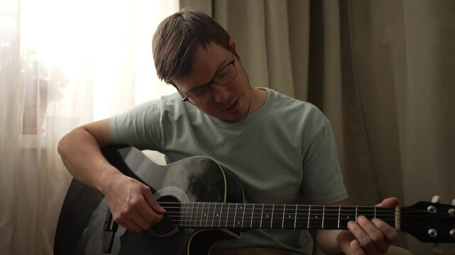 Young man in casual clothing sits and plays an acoustic guitar, practicing chords on a six-string instrument. Natural light filters through a nearby window, enhancing the creative atmosphere
