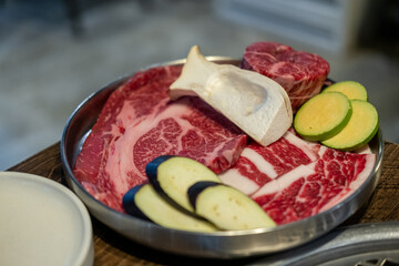 A close-up shot of premium cuts of marbled Hanwoo beef placed on a metal tray, with sliced eggplant and zucchini and eryngii mushroom