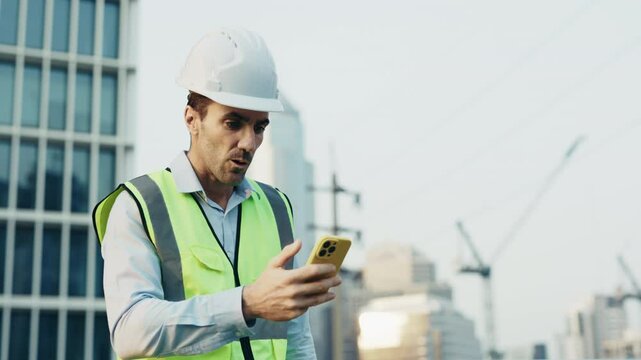 Portrait of man engineer in safety vest showing victory gesture looking at phone against construction site. Male contractor receives notification about successful project update