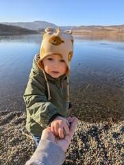 A two-year-old boy eagerly receives stones from his mother’s hands, excited to toss them into the...
