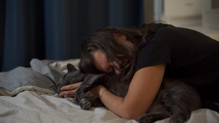 Adult 44 year old woman and gray domestic cat lying on bed in bedroom. Happy woman hugging and kissing her cat, stroking his belly.