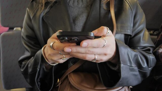 Woman in black leather jacket typing message on smartphone for social media in public transport, close-up. Hands using a chat or messaging app on mobile phone. Searching for information on Internet