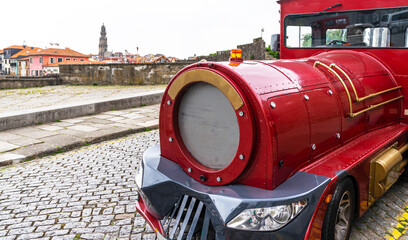 A vibrant red touristic train on the cobbled streets of Porto, with the iconic Clerigos Tower in the background under a soft overcast sky.