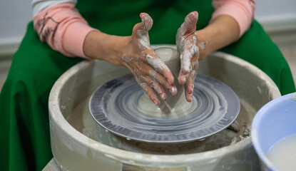 A person wearing a green apron with hands covered in clay is shaping a piece of pottery on a spinning wheel. Fully engaged in the creative process with a focus on the clay piece.