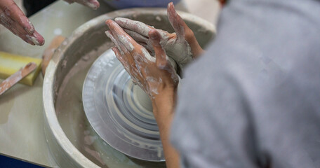 Close-up of a person's hands shaping clay on a pottery wheel. The individual's hands are coated in wet clay, meticulously molding the material. The process takes place in a pottery studio.