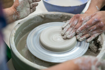 Close-up of hands covered in wet clay as two people collaboratively shape a pottery piece on a spinning wheel, emphasizing the teamwork and artistry involved in ceramic craftsmanship.