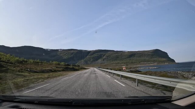 Driving through Norway's tundra on E69 highway. Scenic views of moss-covered rocks, low grass, and the sea on the right, with towering mountains ahead under a crisp, clear sky.