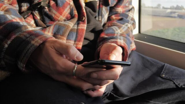 Man in vintage flannel shirt typing message on smartphone for social media in public transport, close-up. Hands using chat or messaging app on mobile phone. Searching for information on Internet.