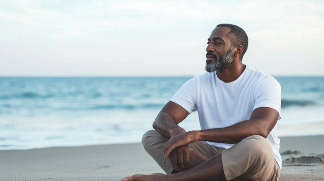 An African American man sits quietly on the beach, his posture relaxed and contemplative, with the vast ocean stretching out before him and ample copy space on either side.