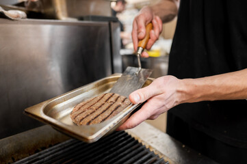 A chef handles a grilled burger patty with a metal spatula, preparing it for serving. The kitchen setting highlights a focus on food preparation and culinary skills.