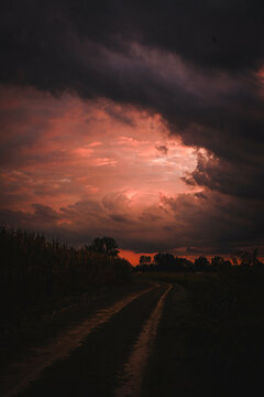 L'immagine mostra un paesaggio rurale al tramonto o all'alba, con un cielo drammatico dai toni rosso-arancio che sfuma nel grigio scuro delle nuvole minacciose. Un sentiero sterrato si snoda tra campi