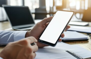 Close-up of a businessman's hand using a white, blank-screen mobile phone with a laptop on the desk in the office. This is a business concept mockup for online social media marketing and digital adver