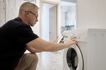 Obraz premium A middle-aged man reads the operating instructions for a washing machine on his smartphone and turns on the washing mode.