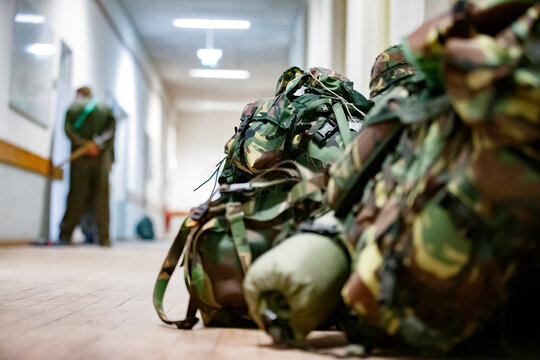 Military backpacks rest on the floor of a barracks hallway while personnel wait in the background during a training session at a military facility