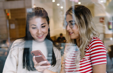 Smiling multiracial female friends sitting in cafeteria on free time watching funny video on smartphone together, positive caucasian girl showing photo on mobile phone to asian best friend on meeting