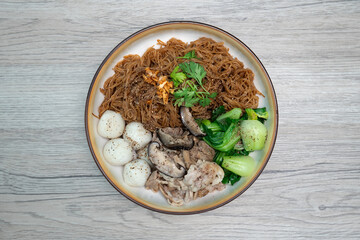 Dry rice vermicelli noodles mixed with dark soy sauce, fish balls, stewed pork and Chinese mustard greens on wooden table, Top view.