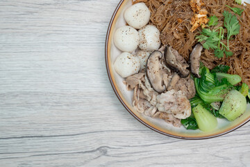 Dry rice vermicelli noodles mixed with dark soy sauce, fish balls, stewed pork and Chinese mustard greens on wooden table, Top view.