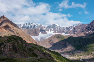 Fototapeta premium Scenic landscape with glacier tongue among rocky ridges and sharp snow-capped mountain range in sunlight under clouds in blue sky. Alpine valley with green hills and rocks against ice and sheer crags.