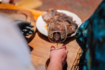 A close-up of a hand holding a glass of red wine, with a plate of roasted meat in the background on a rustic wooden table..